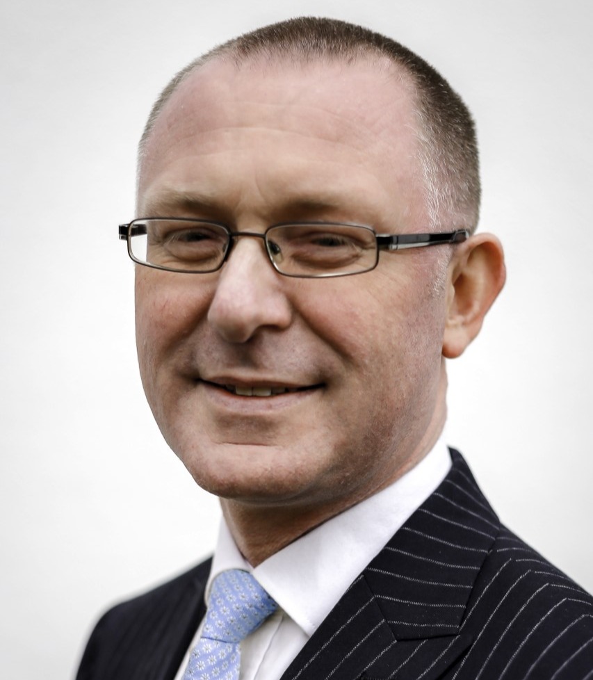 Portrait of a Simon Walker, Senior HR Business Partner, wearing glasses, a dark suit, white shirt, and blue tie, set against a plain white background.